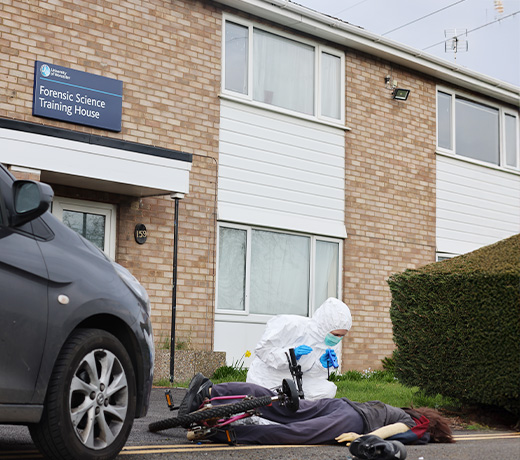 A student in a forensic white suit examining a 'hit and run' victim in front of the Forensic Science Training House