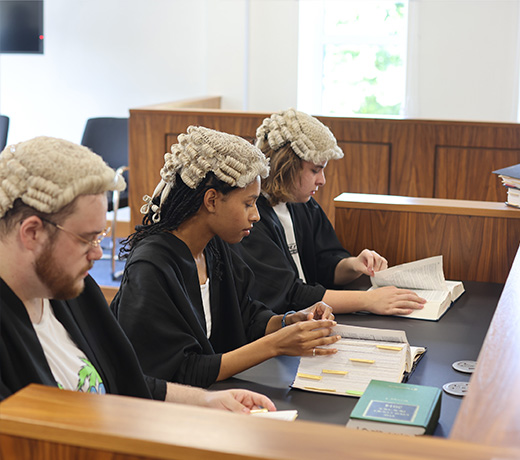 Three Law students wearing robes and wigs seated in the mock court room and looking through papers