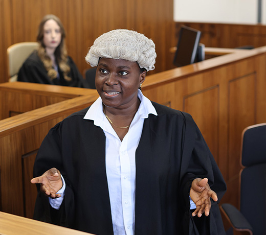 A Law student wearing a wig and robe in the mock courtroom, talking to someone out of view and gesturing with her hands