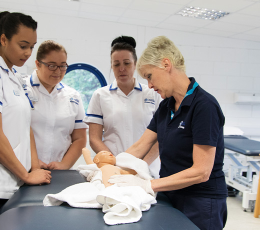 Three students gathered around a Midwifery lecturer demonstrating with a baby manakin