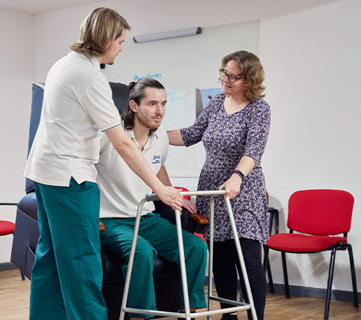 A lecturer demonstrating how to help a patient out of an armchair, with two OT students