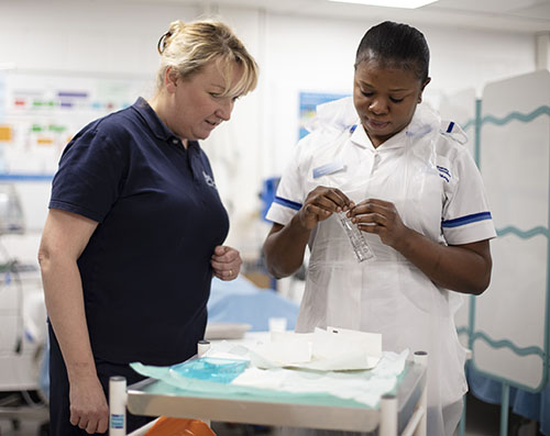 A Nursing lecturer, dressed in a dark blue uniform, is assisting a nursing student, dressed in a white University of Worcester Nursing student uniform, to set up a task on a trolley.