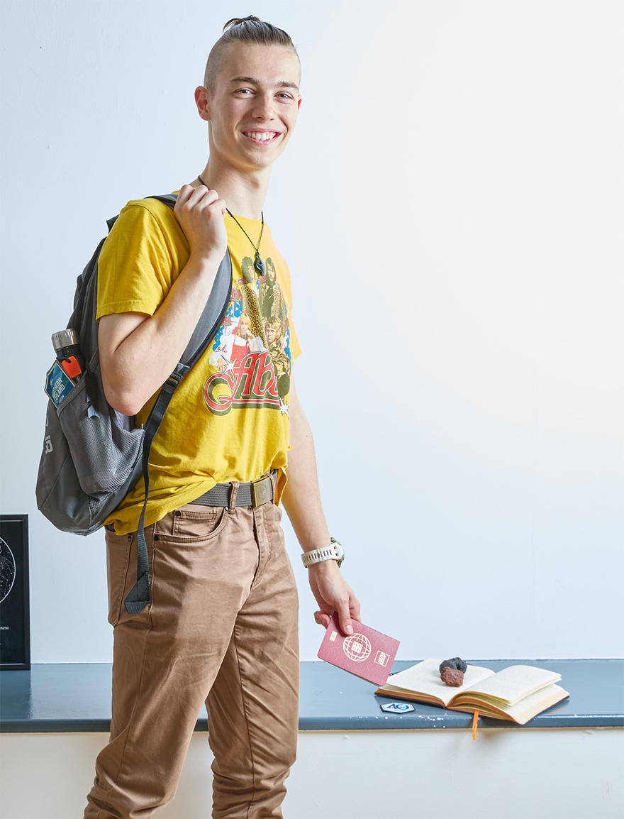 A young person wearing a yellow Abba t-shirt and a backpack standing and holding a passport