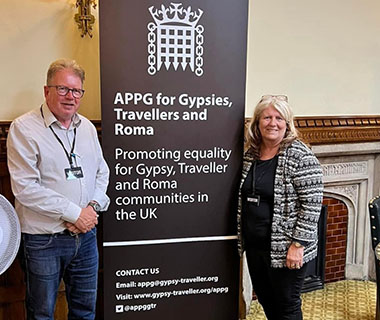 Peter Unwin and Josie O Driscoll are standing next to a sign for the Gypsy Roma Traveller APPG meeting